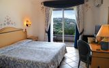 Helles Hotelzimmer mit Doppelbett, Schreibtisch und Balkon – liebevolle florale Details an Wänden und Vorhängen, Blick auf grüne Hügellandschaft.