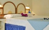 Gemütliches Doppelzimmer mit mediterranem Flair – dekoratives Holzbett mit Sonnenmotiv, frischen Blumen auf dem Bett und Urlaubsaccessoires wie Sonnenbrille und Reiseführer.
