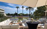 Moderne Lounge-Terrasse mit weißen Sitzgruppen und Sonnenschirmen, Blick auf Palmen und Gartenanlage unter blauem Himmel.