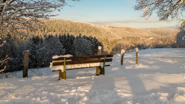 Mit Schnee bedeckte Holzbank in der Eifellandschaft im Winter