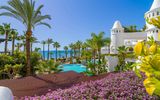 Blick über blühende Pflanzen auf eine paradiesische Hotelanlage mit weißen Türmen, tropischen Palmen, einer großzügigen Poollandschaft und dem strahlend blauen Meer im Hintergrund.