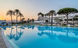 Weitläufiger Hotelpool im Abendlicht mit spiegelnden Palmen, weißen Liegen und Blick auf ein elegantes Resortgebäude vor untergehender Sonne.