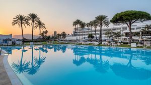 Weitläufiger Hotelpool im Abendlicht mit spiegelnden Palmen, weißen Liegen und Blick auf ein elegantes Resortgebäude vor untergehender Sonne.