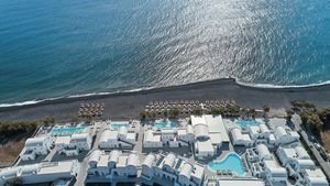 Luftaufnahme des Costa Grand Resort & Spa auf Santorin – elegante Hotelanlage direkt am schwarzen Lavastrand von Kamari mit mehreren Pools, weißen Gebäuden im Kykladenstil und herrlichem Blick auf das funkelnde Meer.