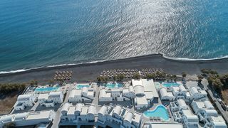 Luftaufnahme des Costa Grand Resort & Spa auf Santorin – elegante Hotelanlage direkt am schwarzen Lavastrand von Kamari mit mehreren Pools, weißen Gebäuden im Kykladenstil und herrlichem Blick auf das funkelnde Meer.