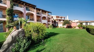 Hotelanlage im mediterranen Stil mit rosa-ockerfarbenen Fassaden, Balkonen und gepflegter Gartenanlage – umgeben von Palmen, Büschen und großen Felsen unter strahlend blauem Himmel.