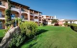 Hotelanlage im mediterranen Stil mit rosa-ockerfarbenen Fassaden, Balkonen und gepflegter Gartenanlage – umgeben von Palmen, Büschen und großen Felsen unter strahlend blauem Himmel.