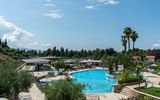 Moderner Hotelpoolbereich mit großer, geschwungener Poolanlage, umgeben von Sonnenliegen, Palmen und mediterraner Vegetation – im Hintergrund flache Gebäude, Pinien und blauer Himmel.