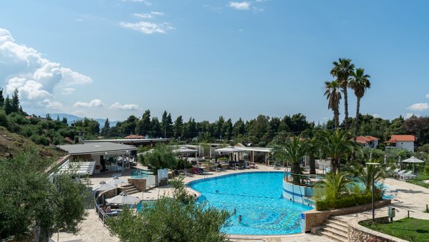 Moderner Hotelpoolbereich mit großer, geschwungener Poolanlage, umgeben von Sonnenliegen, Palmen und mediterraner Vegetation – im Hintergrund flache Gebäude, Pinien und blauer Himmel.