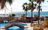 Poollandschaft mit Felsen, Palmen und kleiner Brücke, im Hintergrund Sandstrand mit Sonnenschirmen und Blick auf das glitzernde Meer unter blauem Himmel.