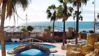 Poollandschaft mit Felsen, Palmen und kleiner Brücke, im Hintergrund Sandstrand mit Sonnenschirmen und Blick auf das glitzernde Meer unter blauem Himmel.