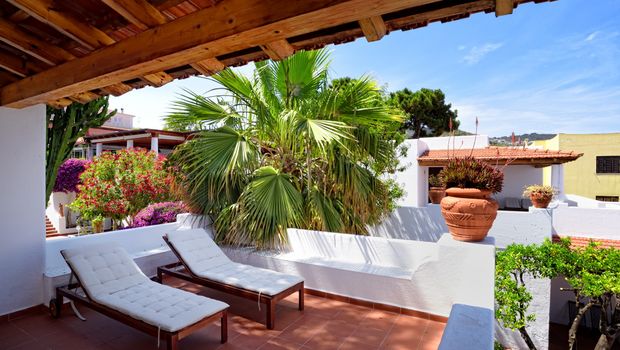 Überdachte Terrasse mit zwei gepolsterten Sonnenliegen, umgeben von Palmen, blühenden Pflanzen und weiß verputzten Mauern – mediterrane Ruheoase mit traditionellem Flair.