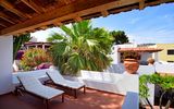 Überdachte Terrasse mit zwei gepolsterten Sonnenliegen, umgeben von Palmen, blühenden Pflanzen und weiß verputzten Mauern – mediterrane Ruheoase mit traditionellem Flair.