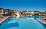 Weitläufige Poollandschaft eines mediterranen Resorts mit Sonnenliegen und Sonnenschirmen – dahinter bunte, terrakottafarbene Hotelgebäude mit Balkonen und üppiger Vegetation unter strahlend blauem Himmel.