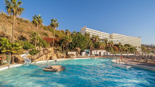 Weitläufige Poolanlage mit Wasserrutsche, Felsenlandschaft und Liegestühlen, umgeben von tropischen Palmen und blühender Vegetation, im Hintergrund ein großes Hotelgebäude an einem sonnigen Hang.