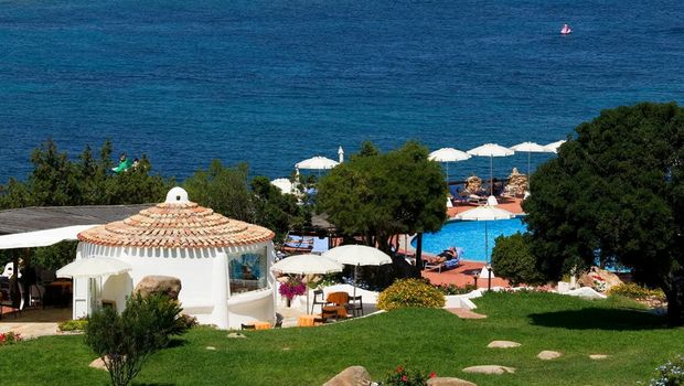 Blick auf einen idyllischen Hotelgarten mit mediterranen Pflanzen, Poolbereich mit Sonnenliegen und weißen Sonnenschirmen – im Hintergrund das tiefblaue Meer, das zum Träumen einlädt.