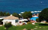 Blick auf einen idyllischen Hotelgarten mit mediterranen Pflanzen, Poolbereich mit Sonnenliegen und weißen Sonnenschirmen – im Hintergrund das tiefblaue Meer, das zum Träumen einlädt.