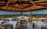 Elegantes Restaurant mit Holzdach und Panoramafenstern – gedeckte Tische mit Blick auf das Meer, grüne Landschaften und ferne Inseln unter blauem Himmel.