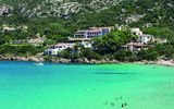 Idyllische Bucht mit kristallklarem, türkisblauem Wasser – im Hintergrund eingebettet in grüne Hügel liegen charmante weiße Hotelgebäude mit roten Dächern, während Badegäste das Meer genießen.