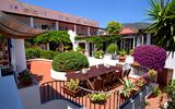 Charmanter Innenhof mit üppiger Bepflanzung, weiß verputzten Wänden und großer Terrasse mit Holzsitzgruppe – mediterranes Flair in ruhiger Umgebung mit Blick auf grüne Hügel.